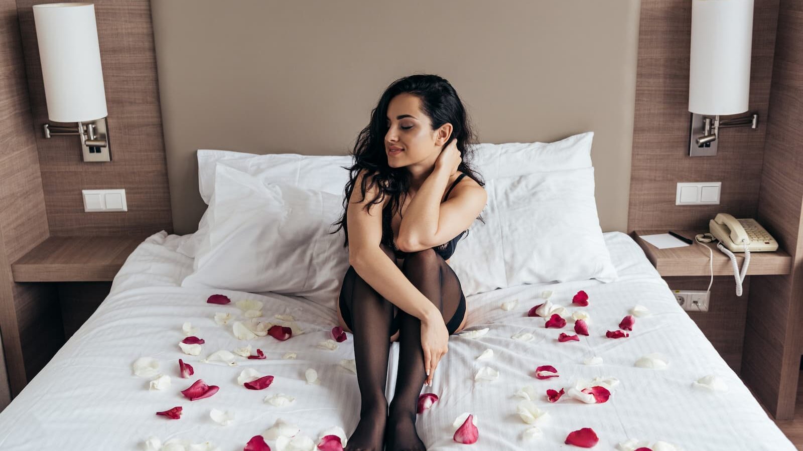 Sexy brunette girl in black stockings sitting on bed with rose petals.